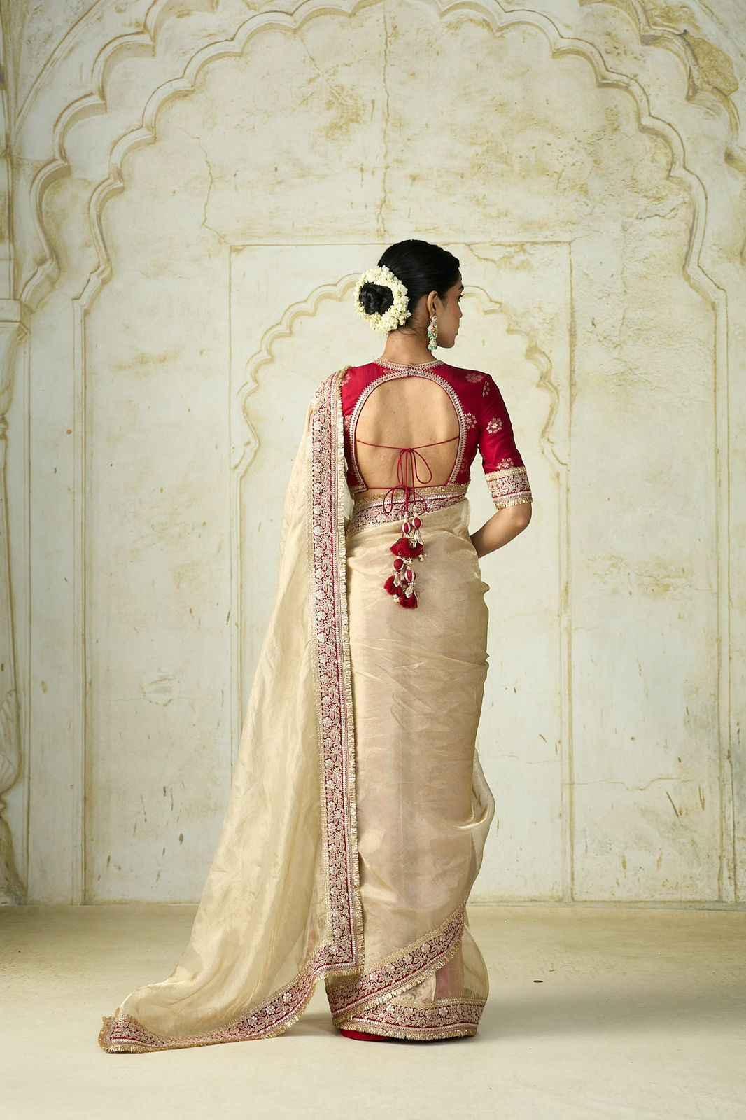 Woman wearing beige silk saree with intricate pink embroidery and red blouse with open back, standing by ornate stone wall