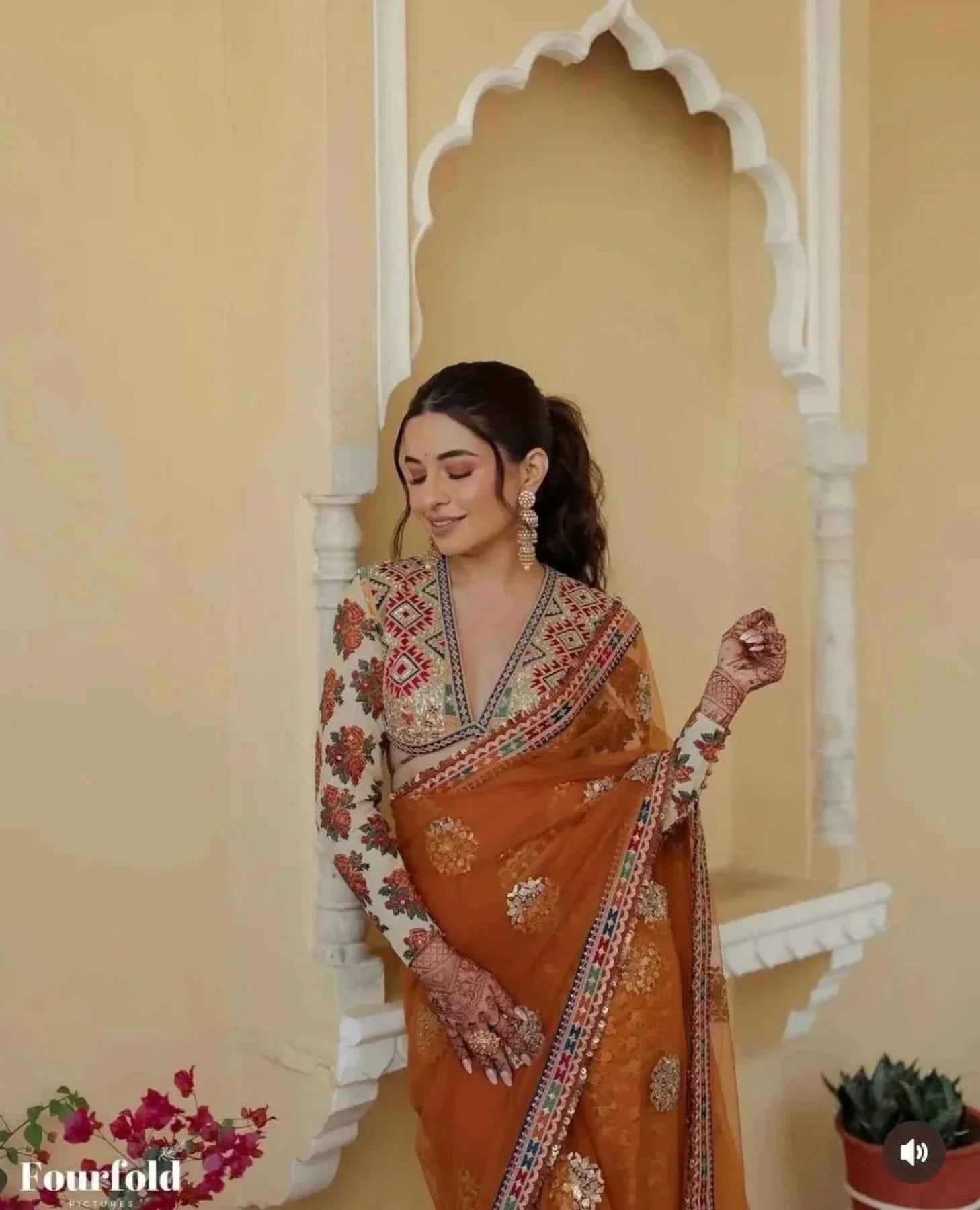 Woman wearing orange net saree with floral printed embroidered blouse standing near traditional arched wall