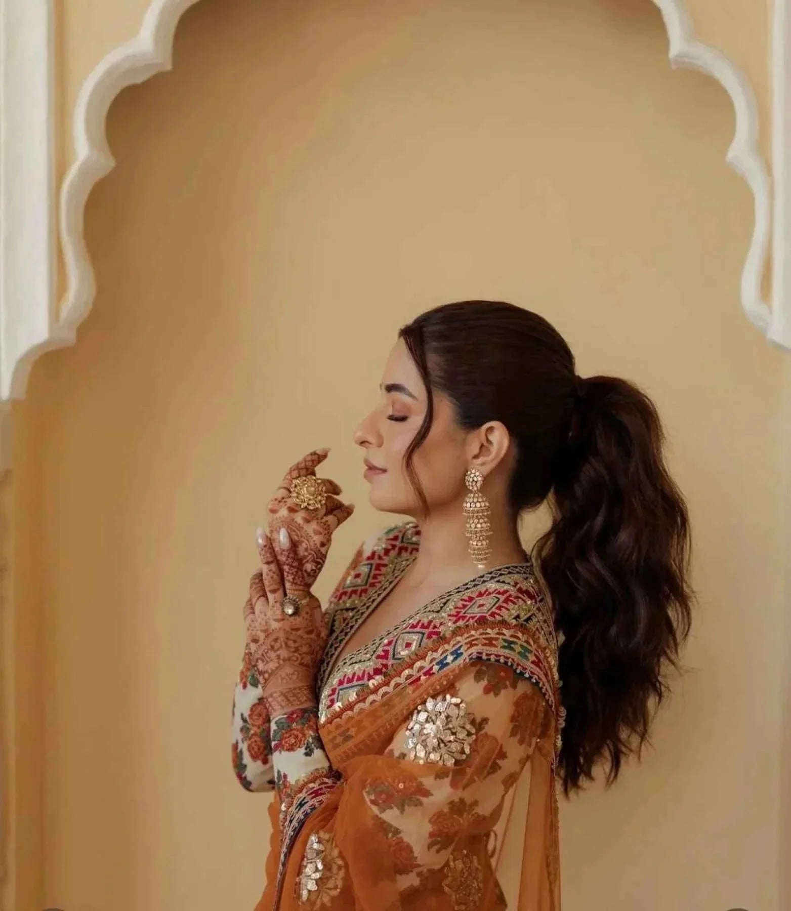 Woman in orange net saree with detailed embroidery and stitched designer blouse, wearing gold earrings and mehndi