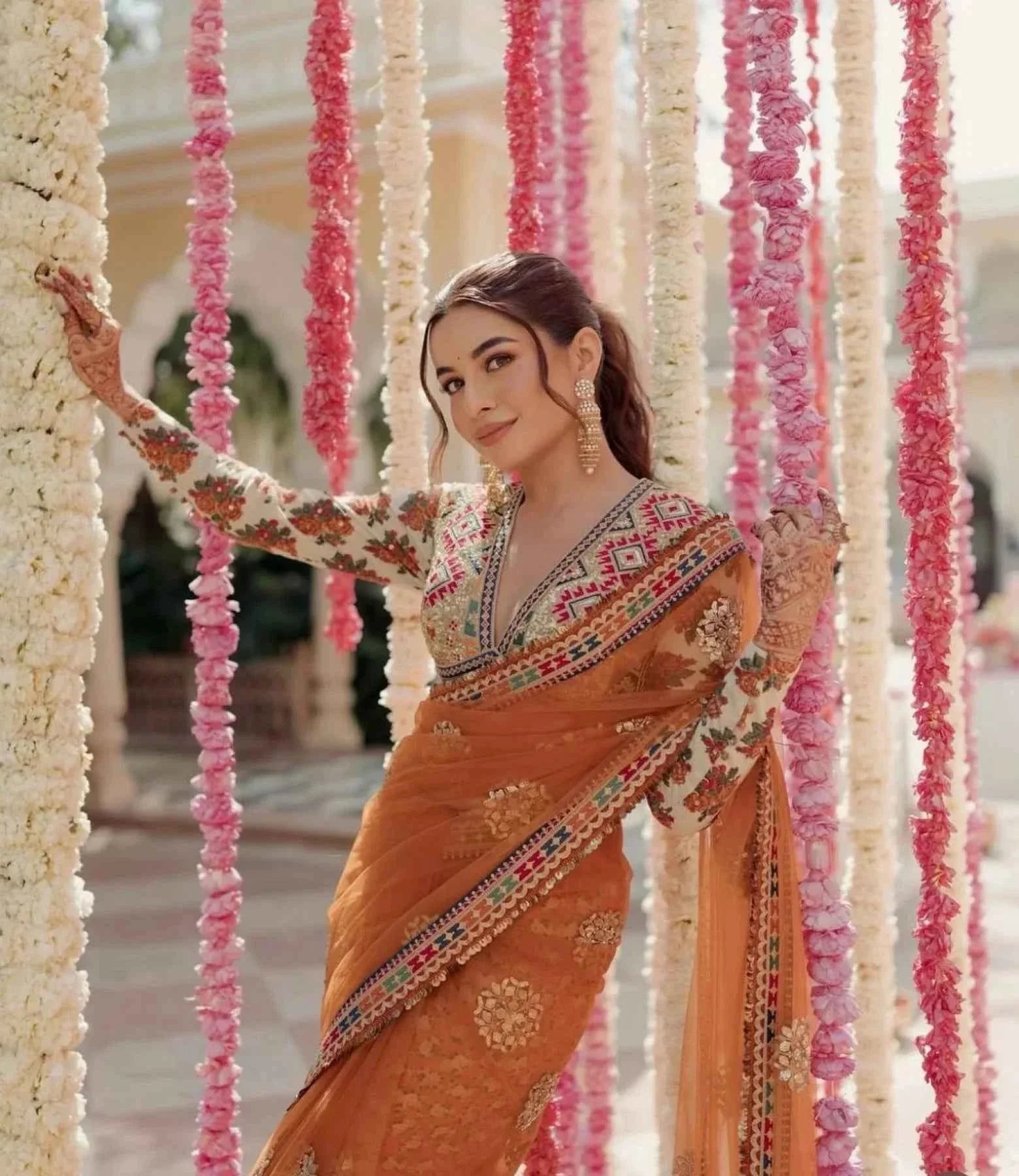 Woman in orange net saree with stitched designer blouse posing among pink and white flower garlands