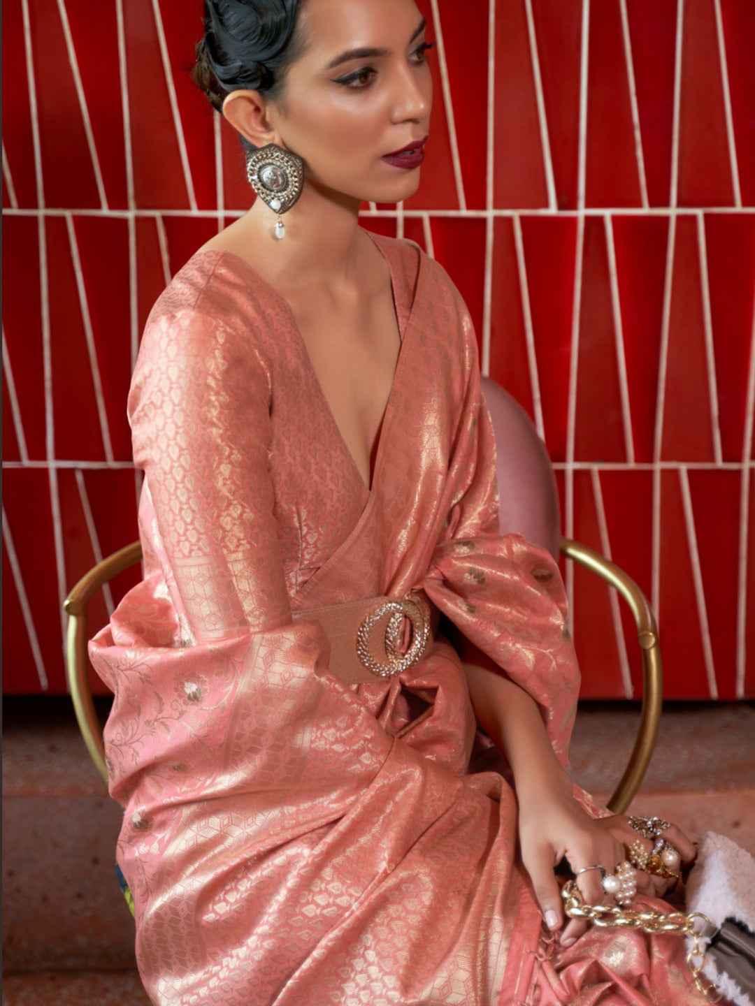 Woman wearing peach silk saree with intricate zari pattern and statement earrings seated against red tiled wall
