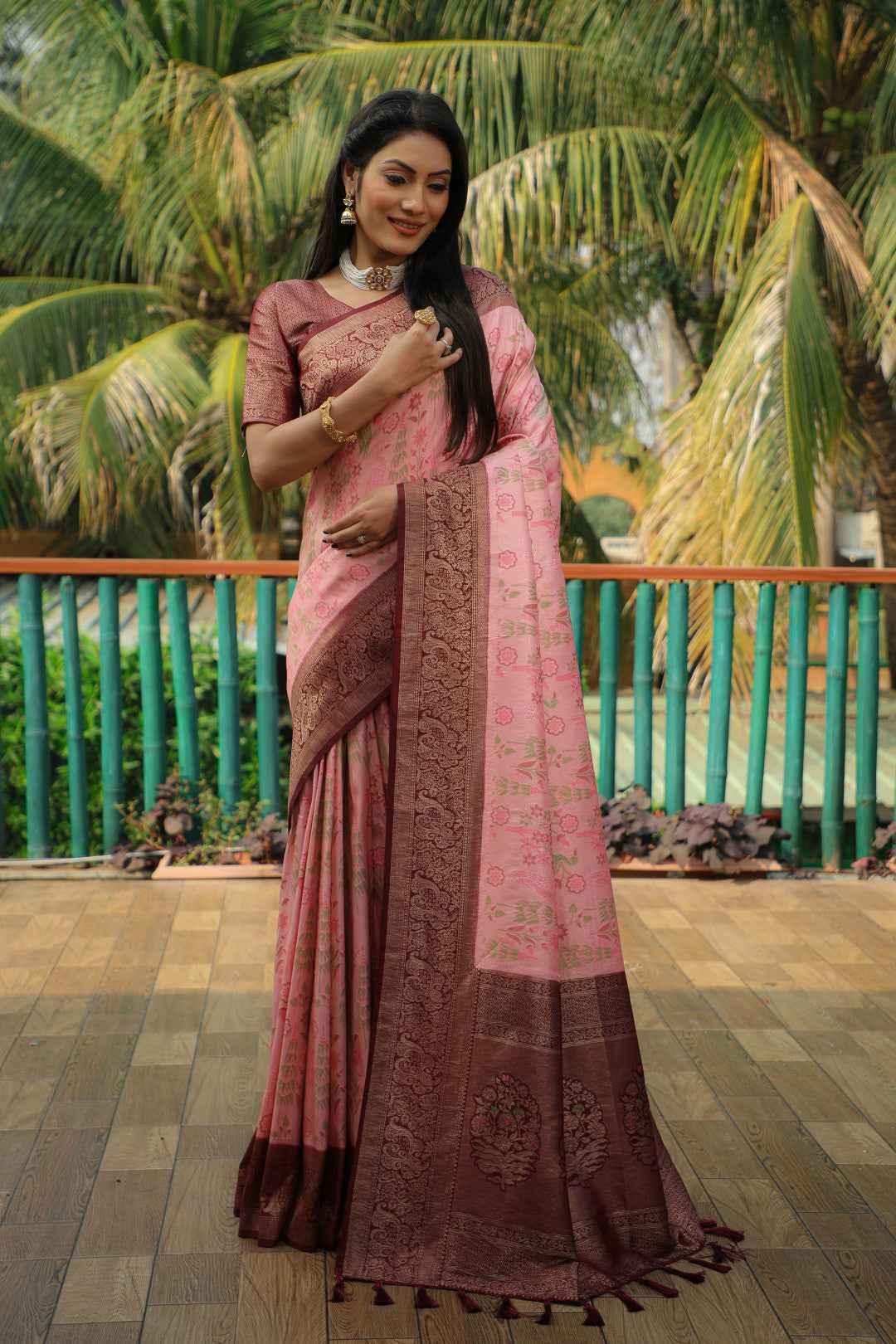 Woman in pink Kanjivaram silk saree with intricate brown zari border, standing on wooden floor near green bamboo railing and palm trees