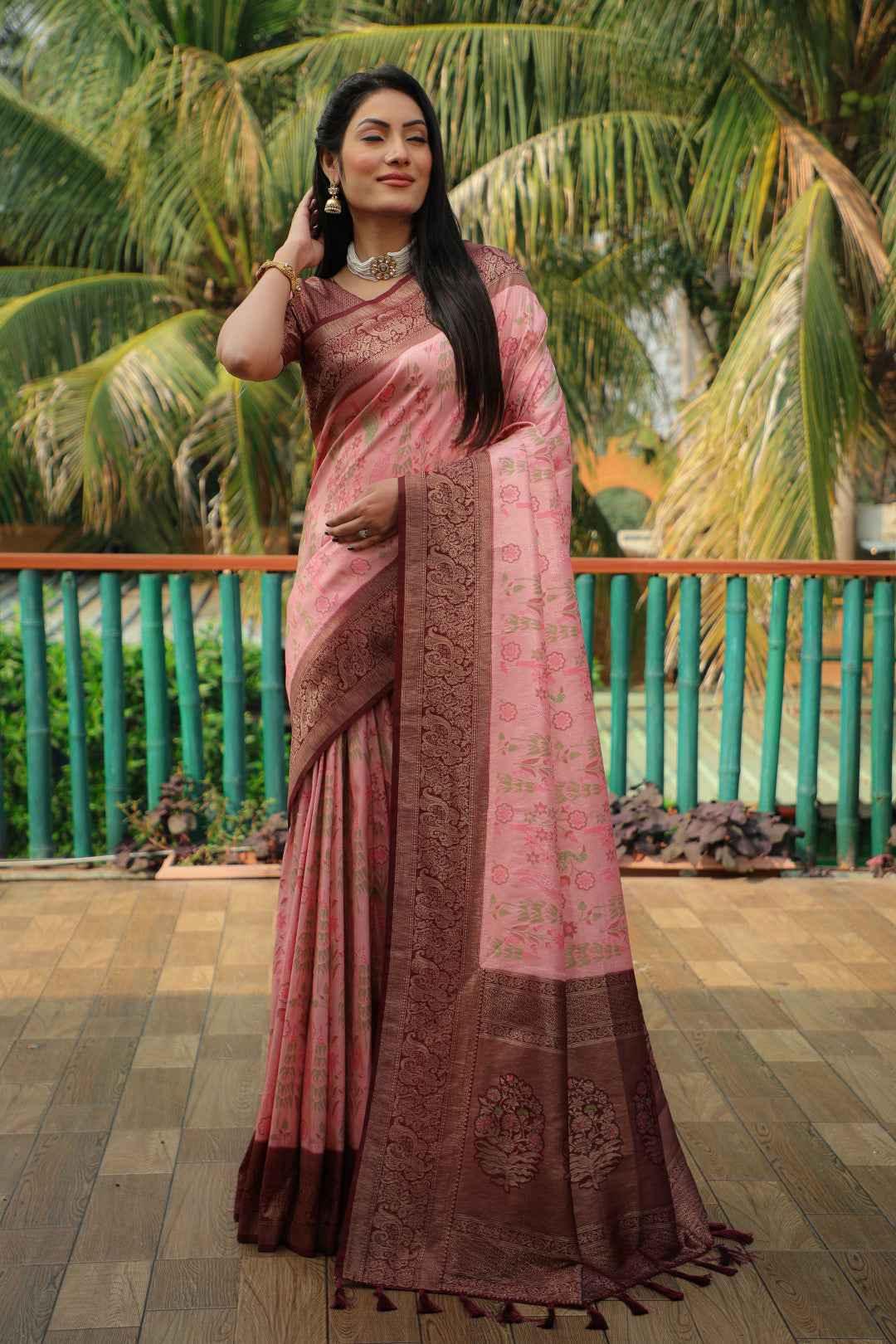 Woman in traditional pink and brown Kanjivaram silk saree with zari weaving, standing on wooden deck with palm trees