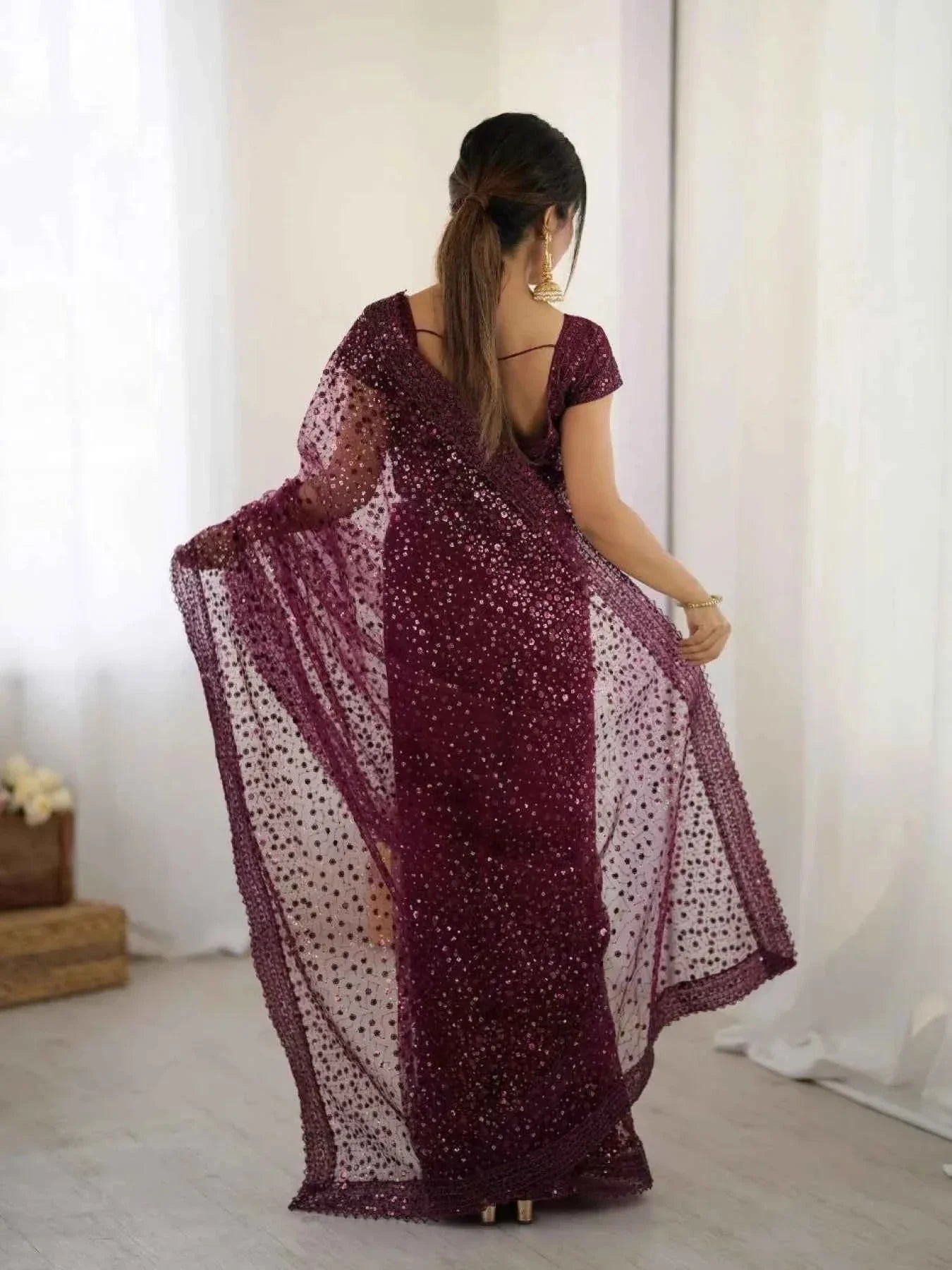 Woman wearing wine net saree with all-over sequin dori embroidery, posing indoors with back view
