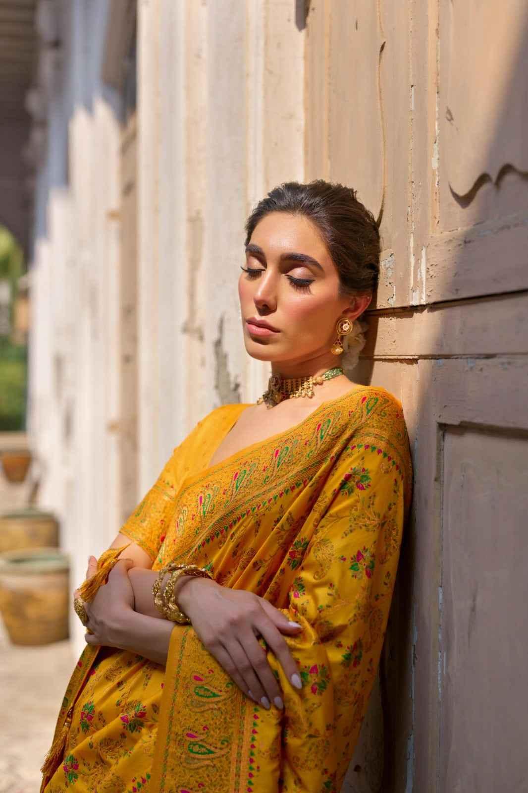 Woman in yellow pashmina silk saree with Kashmiri jal weaving, golden jewelry, leaning against wall